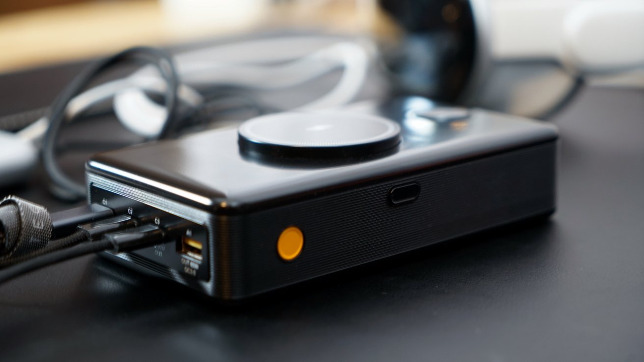 Black electronic device with multiple connected cables, a round top button, and a yellow side button, placed on a dark surface.