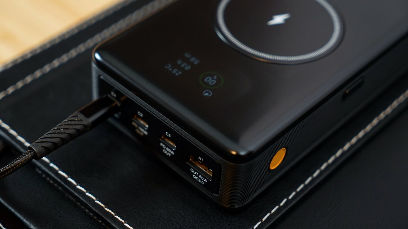 Black portable power bank with multiple USB ports, a glowing power button, and a wireless charging pad on top, placed on a black surface with white stitching.
