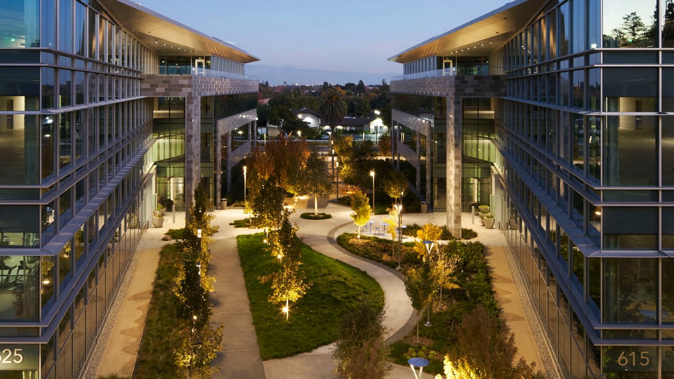Modern office buildings with glass facades flank a landscaped courtyard featuring illuminated pathways, trees, and benches, creating an inviting atmosphere at dusk.