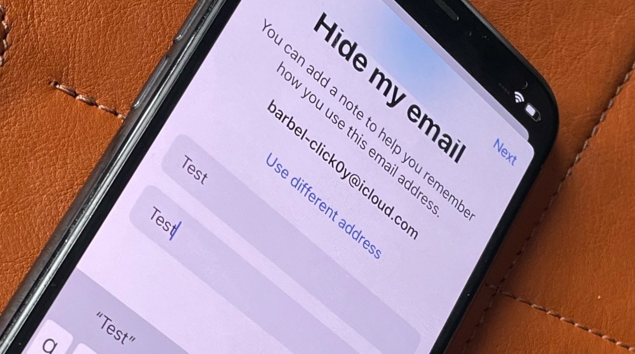 Smartphone screen displaying the 'Hide my email' feature with a test email and note entry fields, on a brown leather surface.
