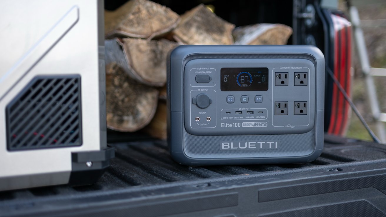Portable power station with display and multiple outlets sits on the back of a vehicle, with stacked firewood in the background.