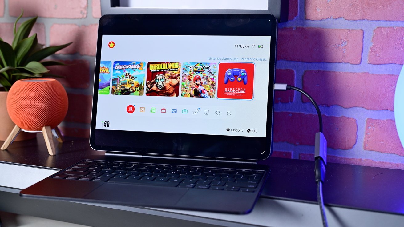Tablet with a Nintendo game interface on a stand connected to a keyboard, on a table with a small orange speaker and potted plant, brick wall background.