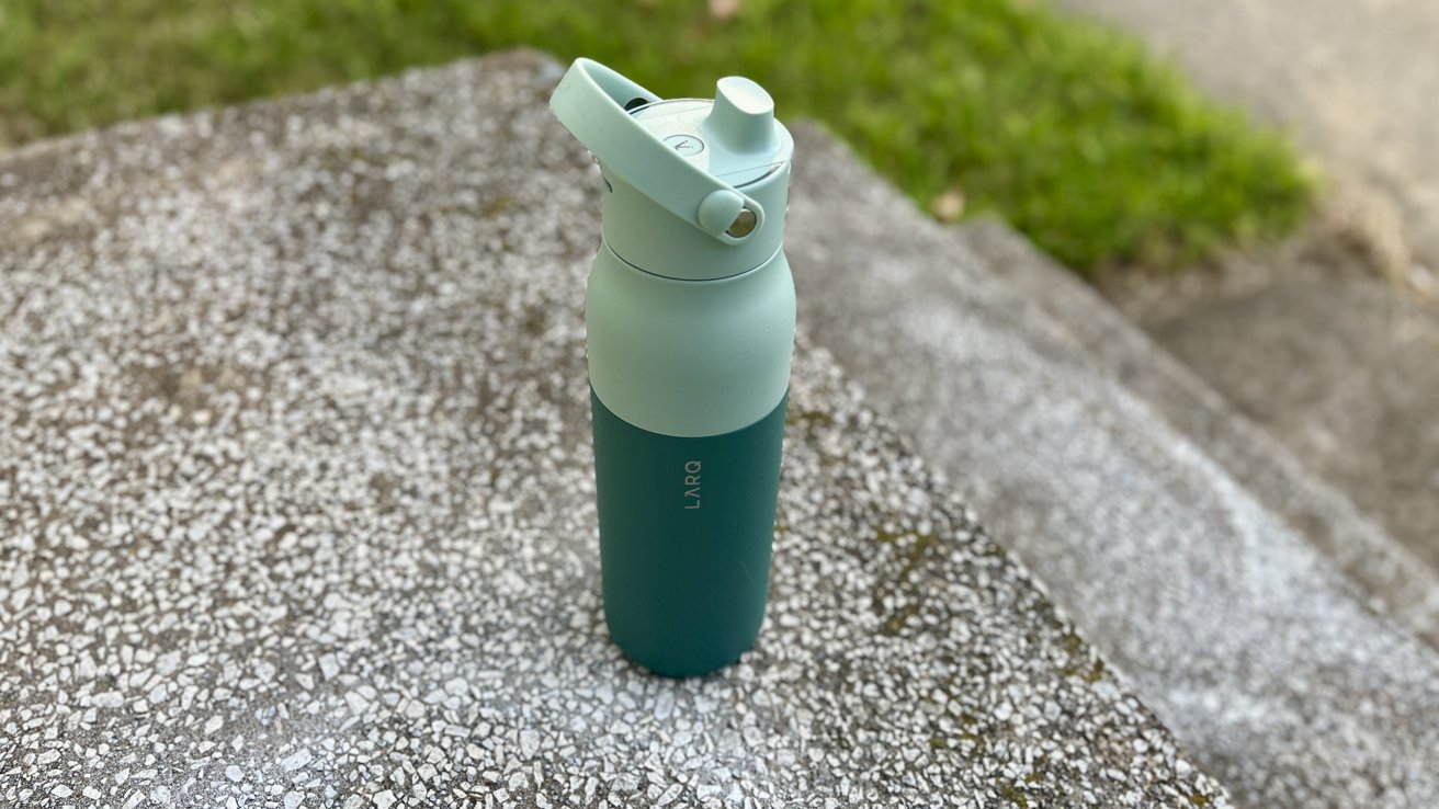 Reusable water bottle with teal and mint colors, flip-top lid, placed on a textured concrete surface outdoors, with grass visible in the background.