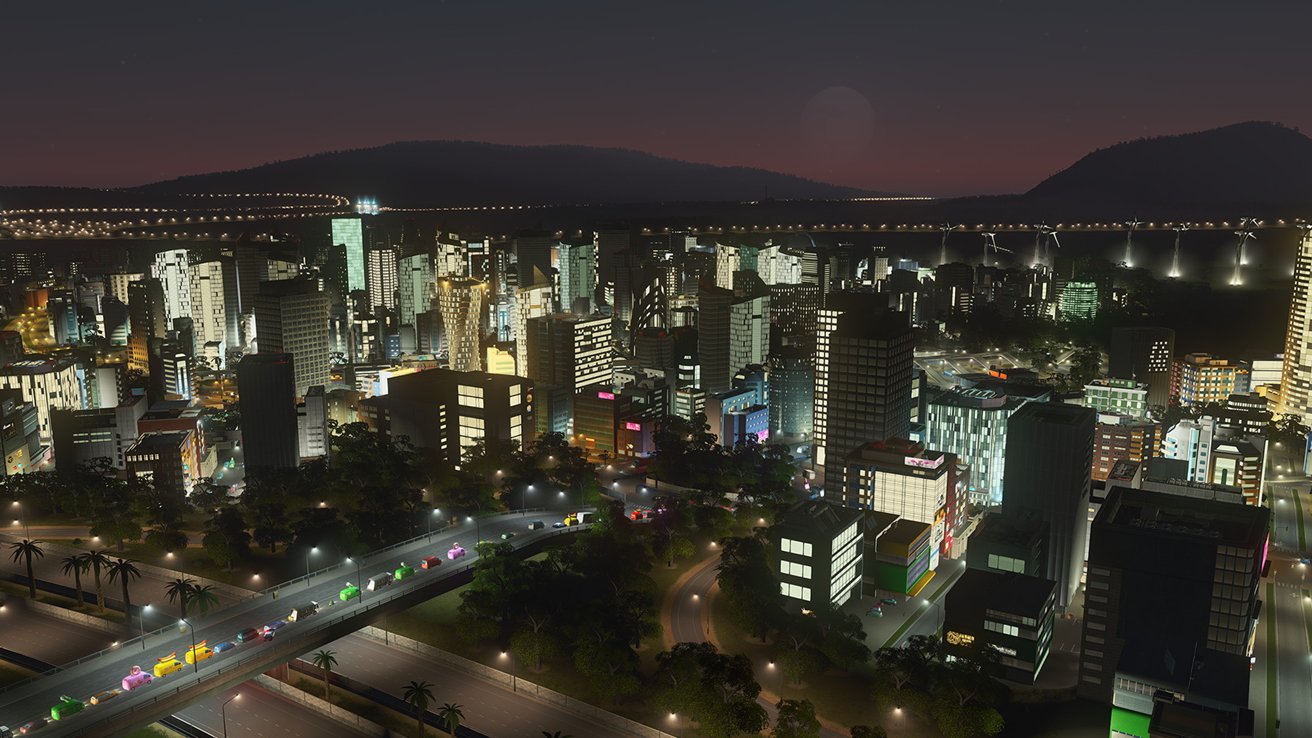 City skyline at night with illuminated buildings, busy roads, and colorful vehicles. Hills and a bridge with lights in the background under a dusky sky.