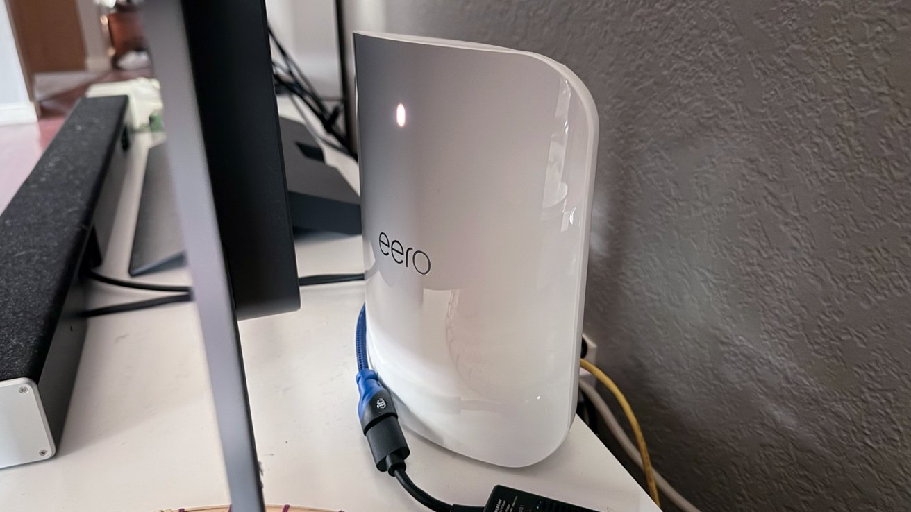 White eero Wi-Fi router with glowing indicator, connected by cable on a desk near a computer monitor and textured wall.