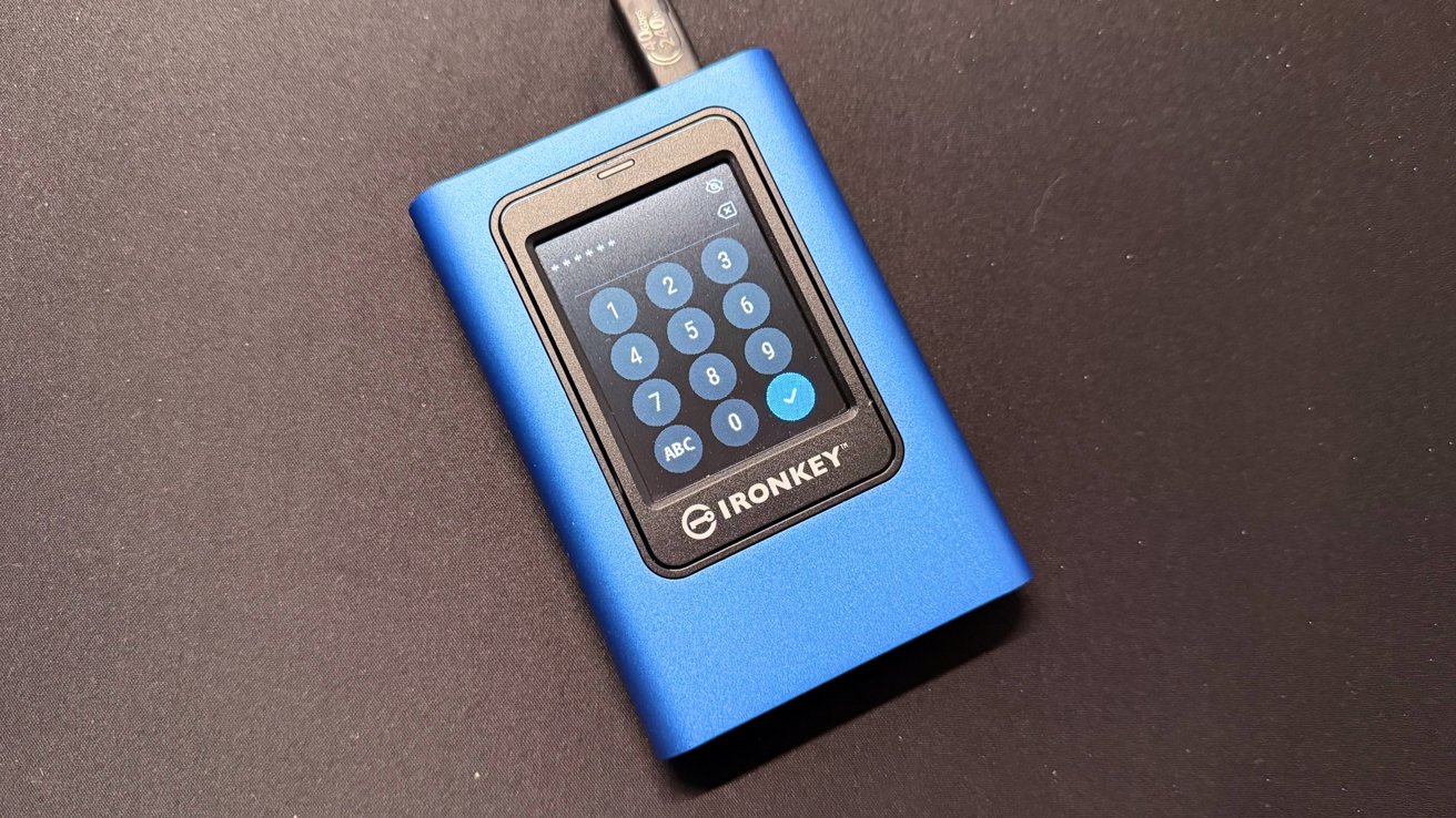 Blue electronic device with a digital keypad on a dark surface. The screen displays numbers, with 'IronKey' branding below. A cable connects to the top.