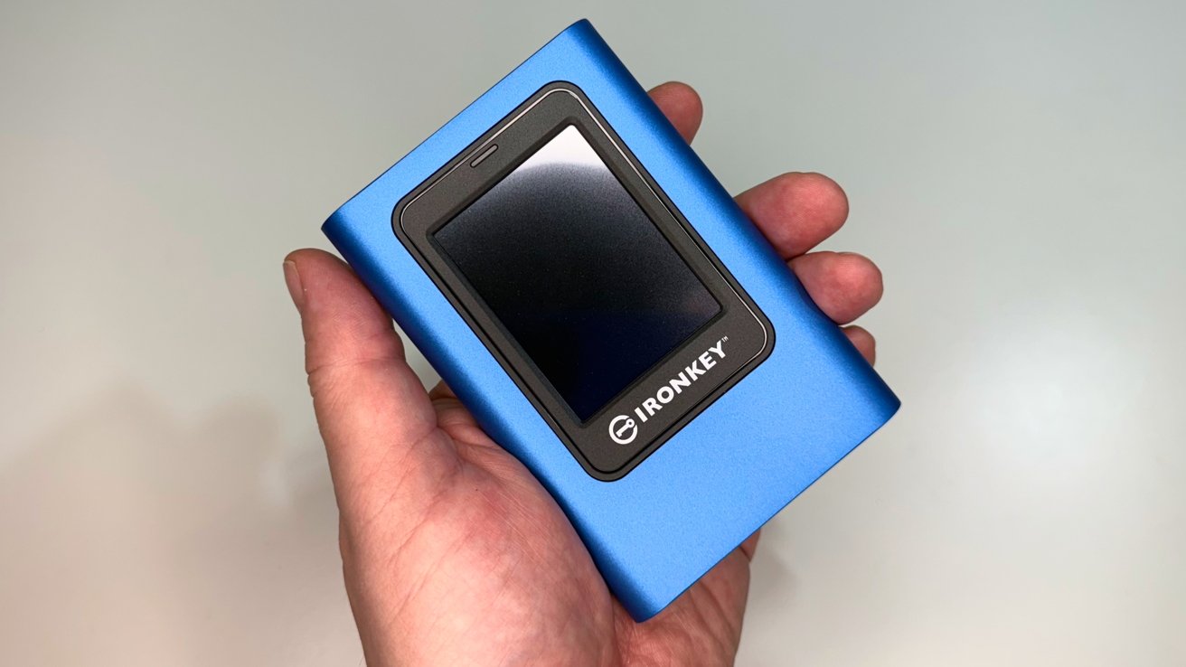 Hand holding a blue electronic device with a screen, labeled IronKey, on a light background.