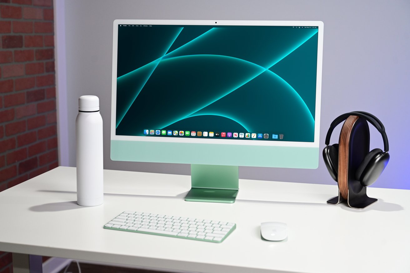 A modern desktop with a sleek computer, wireless keyboard, mouse, white water bottle, and headphones on a stand against a brick and grey wall background.