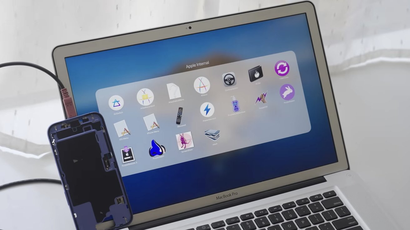 Laptop displaying Apple internal software folder on screen, phone with removed case connected via cable, placed upright against keyboard, white curtain background.