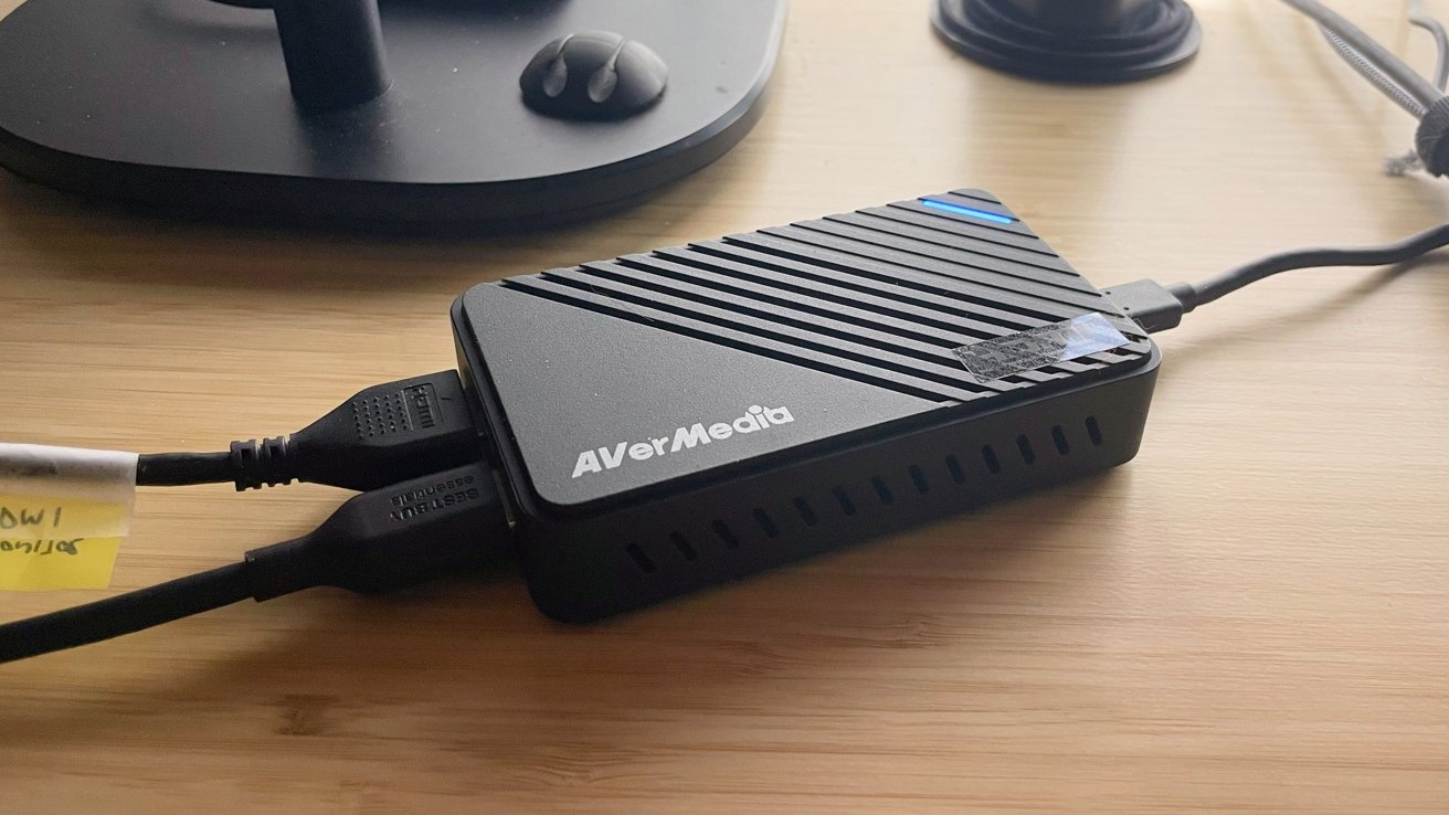A black AVerMedia device with cables attached, placed on a wooden desk. A computer mouse is in the background.