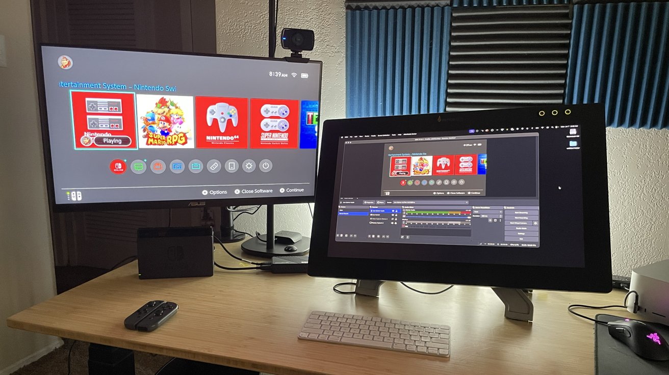 Two monitors on a desk display a Nintendo Switch interface and video editing software. A keyboard, mouse, and game controller are also present. Acoustic foam panels adorn the wall.