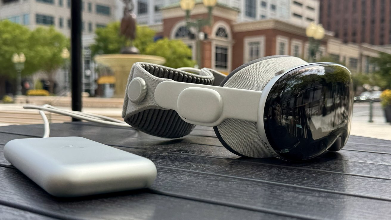 VR headset and charging pack resting on outdoor table in urban setting with blurred buildings and greenery in the background.