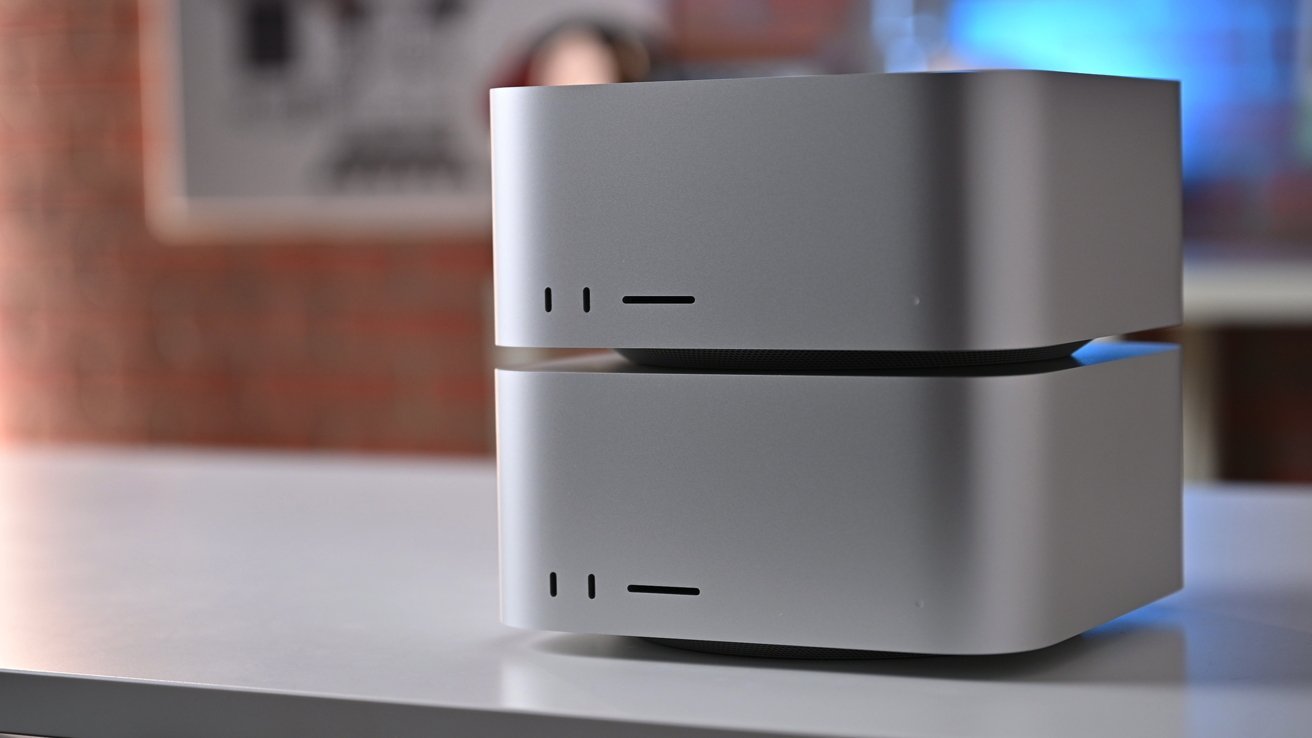 Two stacked, metallic gray computer units with minimalistic design, small ventilation holes, and clean edges placed on a reflective surface against a blurred background.