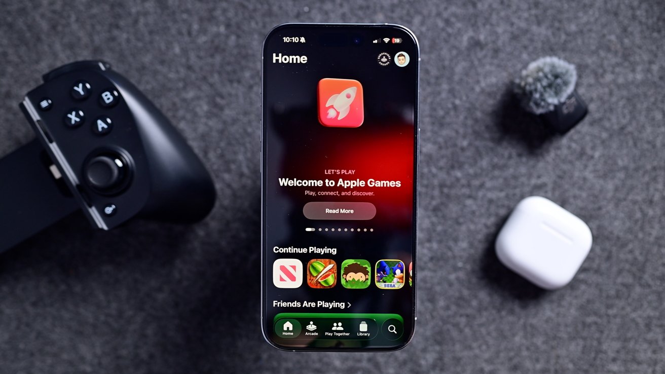 Smartphone displaying Apple Games app, surrounded by a gaming controller, white earbuds case, and microphone on a grey textured surface.
