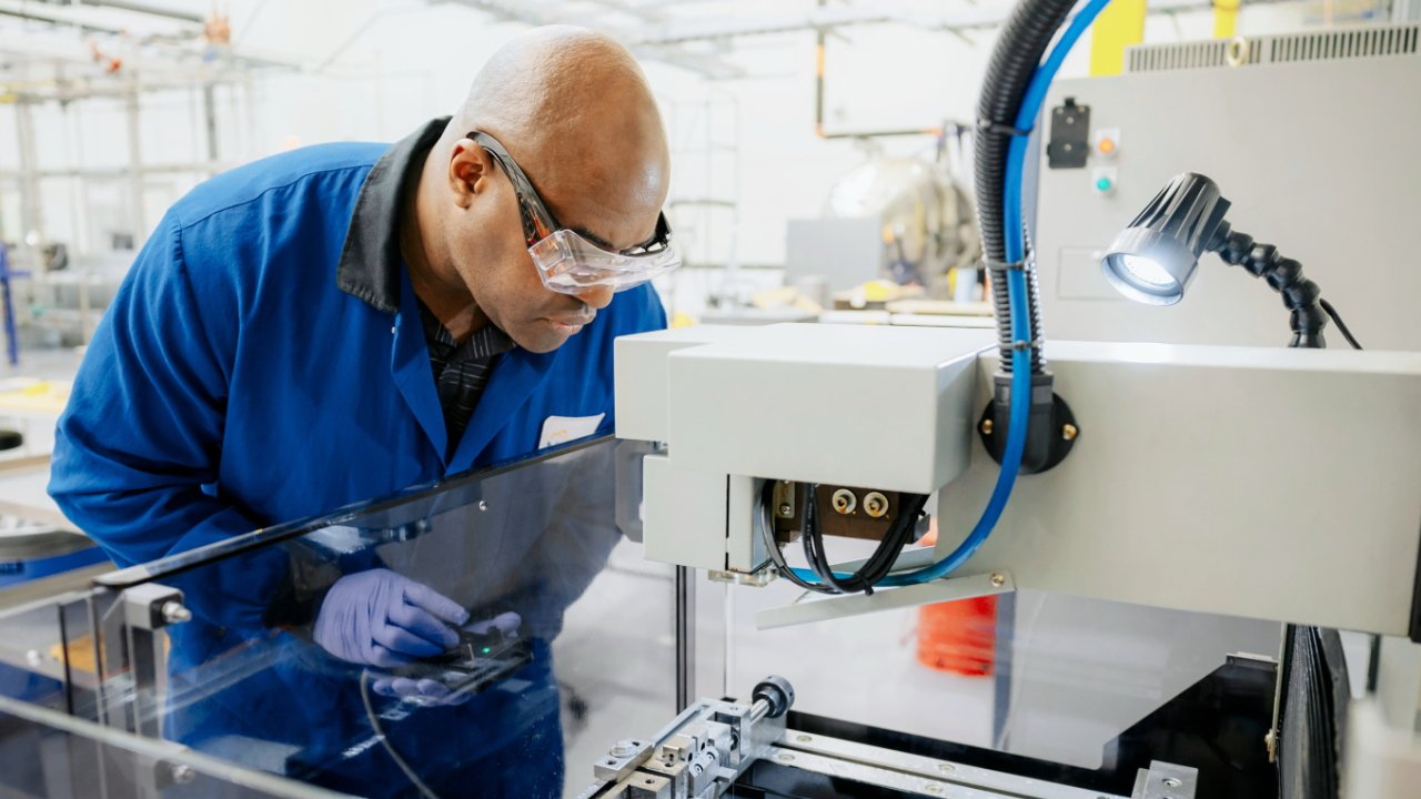 Person wearing safety goggles and blue coat examines machinery under bright light in a lab or industrial setting.