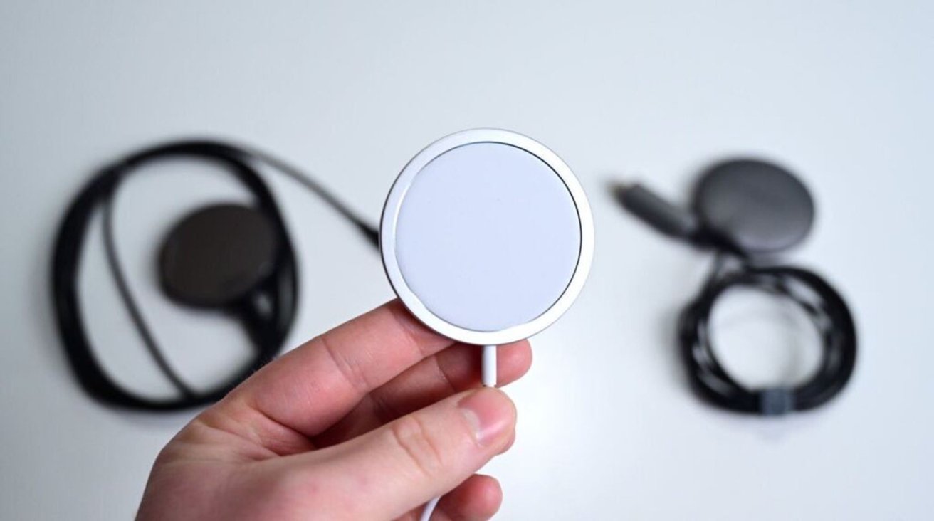 Hand holding a round, white wireless charger, with two similar chargers and cables in the blurred background.