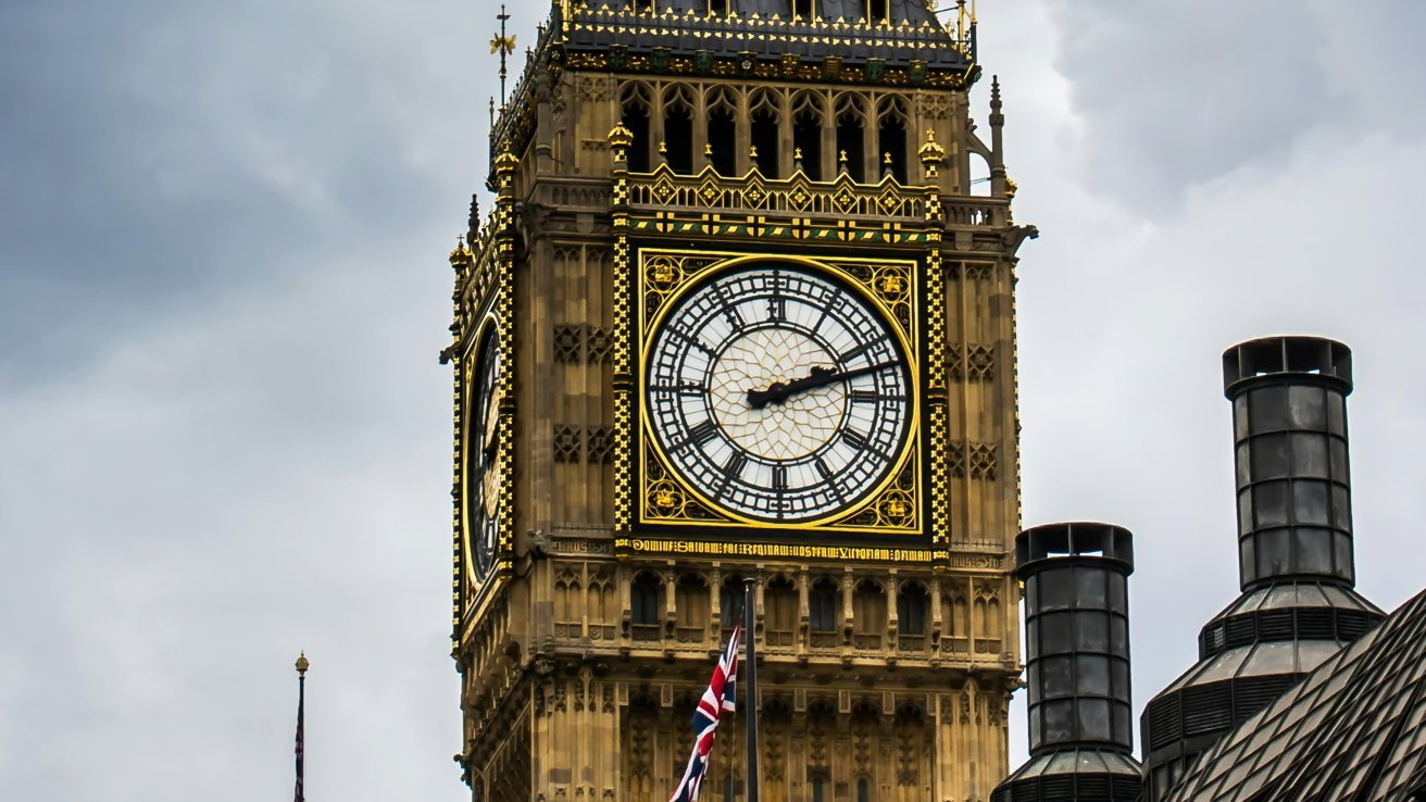 Clock tower with ornate architecture, large white clock face, and golden accents; gray sky background. British flag visible below.