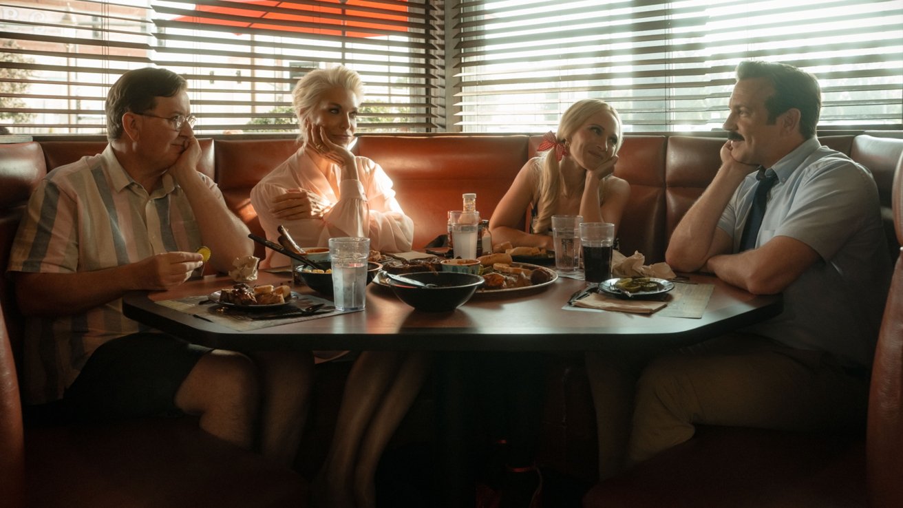 Four people sitting in a diner booth, chatting and enjoying a meal together. Plates of food, glasses, and casual, relaxed atmosphere with warm sunlight filtering through blinds.