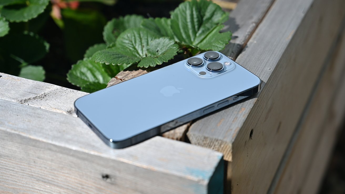 A silver smartphone with triple lenses on a wooden surface beside green leaves.