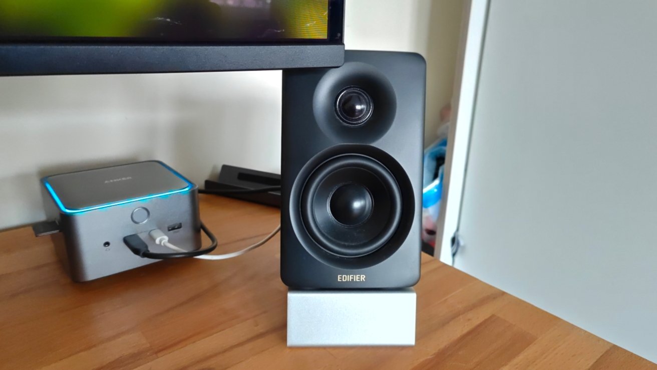 A black speaker stands on a wooden desk, next to a small tech device with a blue-lit edge.
