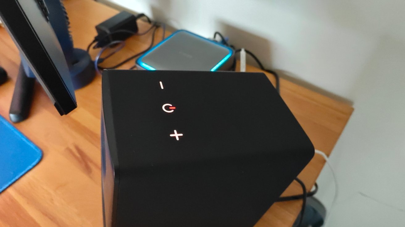 Black electronic device with illuminated touch controls displaying 'I', a power symbol, and '+'. Nearby are a monitor and devices on a wooden surface.