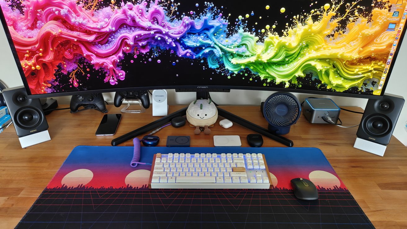 A colorful curved monitor displays vibrant swirls. A desk holds speakers, gaming controllers, a fan, phone, keyboard, mouse, and various small gadgets, with a plush toy in the center.