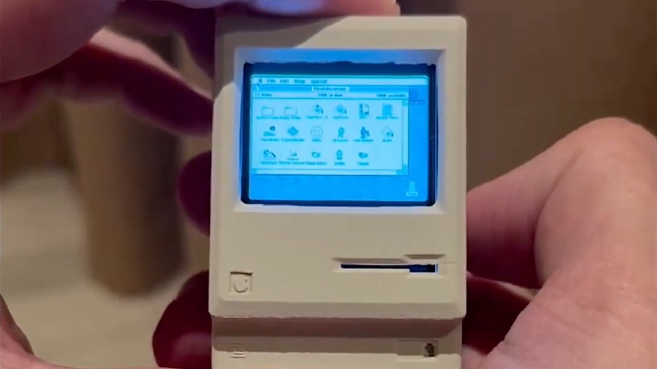 A hand holds a miniature beige computer with a glowing blue screen displaying icons and text, resembling a vintage Macintosh interface.