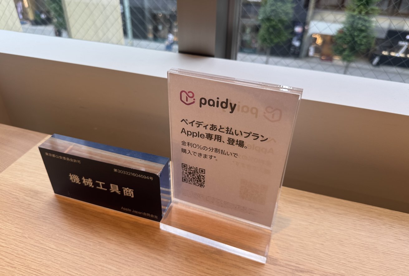 Two sign stands on a wooden table, one with Japanese text and a QR code. A window background shows a street view.
