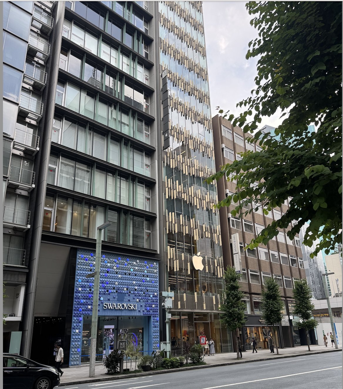 Tall buildings line a city street, including a Swarovski store with a blue facade and an adjacent building with a prominent apple logo. Lush green trees are visible.
