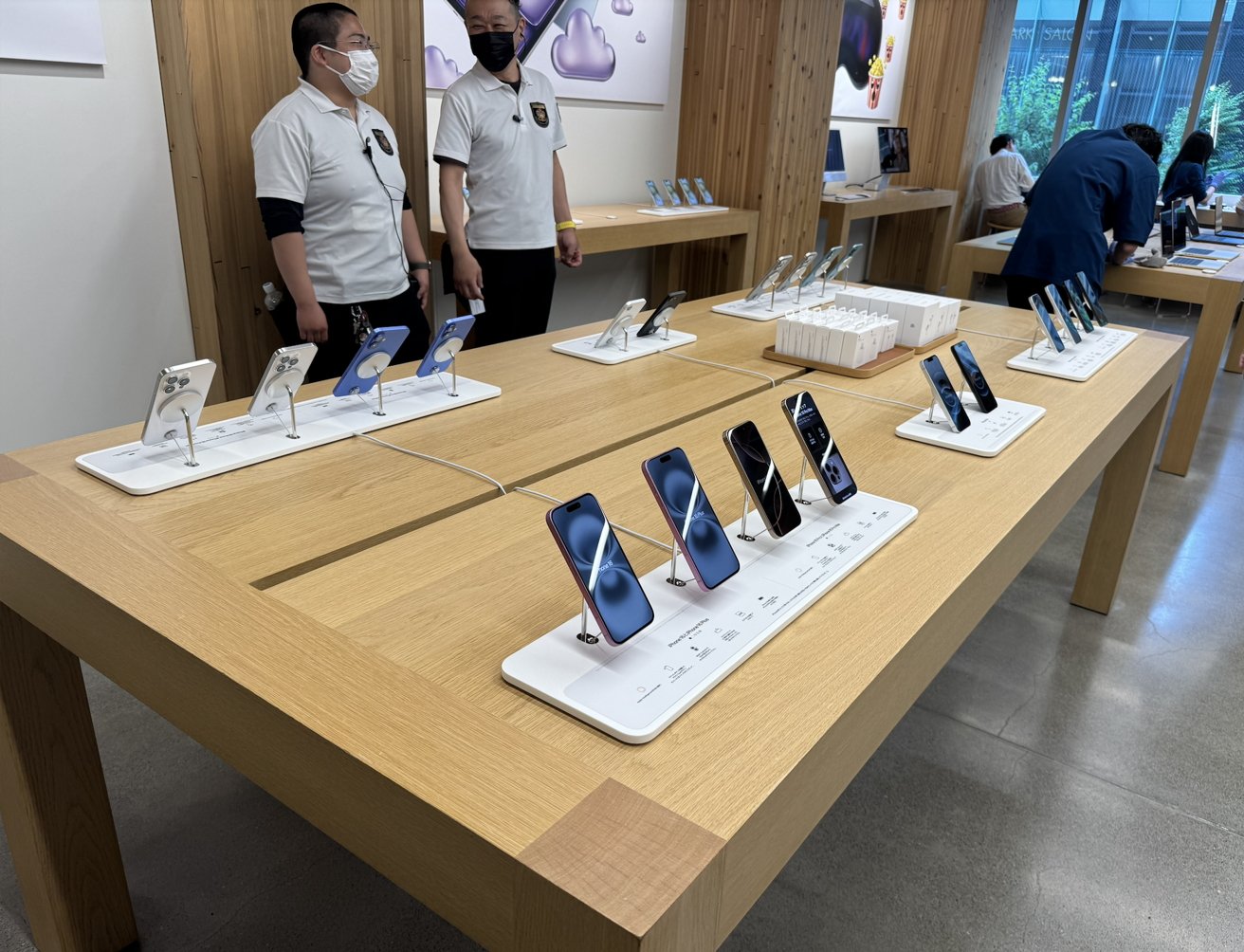 Store display with smartphones on wooden tables, two staff members and customers browsing technology products in a modern retail environment.
