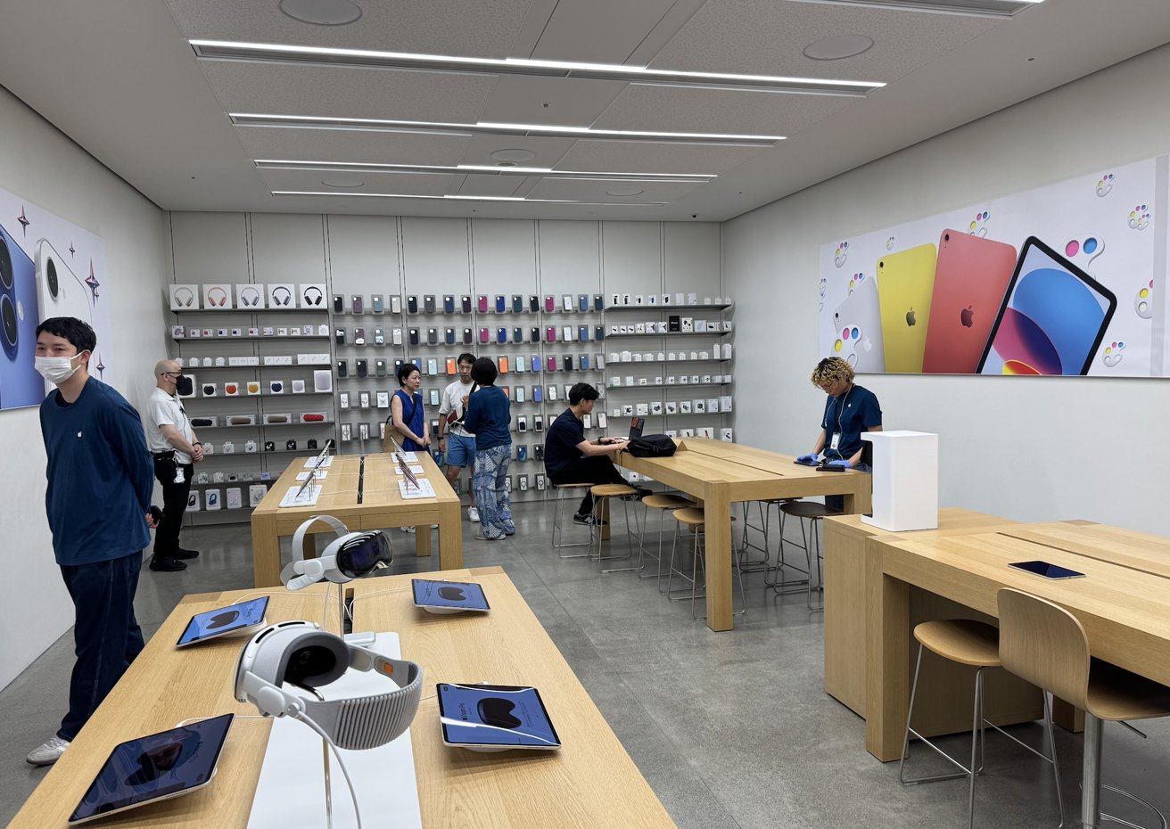 Store interior with wooden tables, electronic devices, and people browsing. Shelves display phone cases. Bright banners feature colorful devices. Staff assist customers.