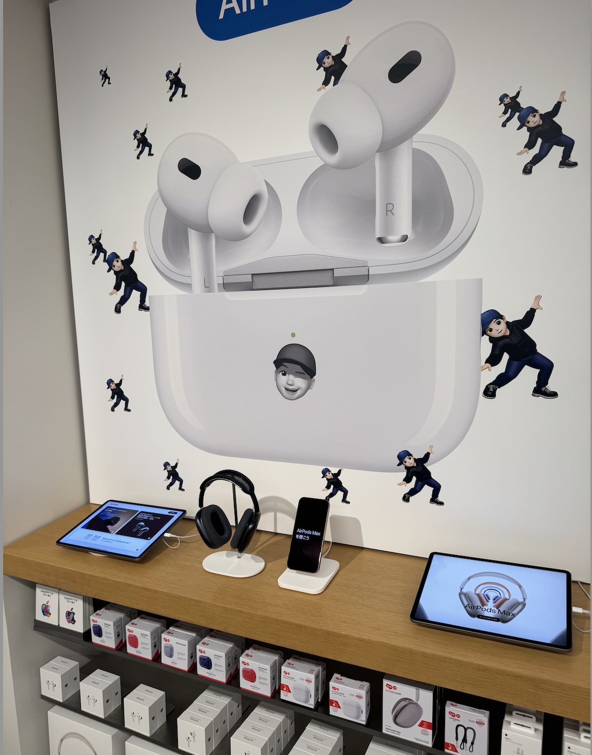 Store display with large AirPods poster above a wooden shelf featuring headphones, a smartphone, tablets, and boxed products. Playful animated characters are scattered throughout the poster.