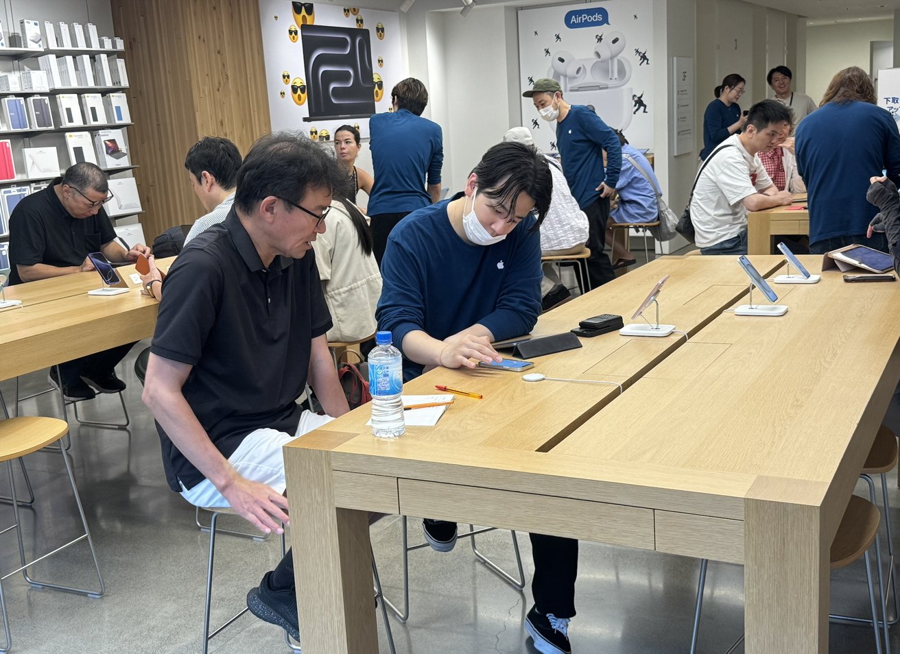 People sitting and interacting with devices in a modern tech store, surrounded by tables, display products, and promotional posters.