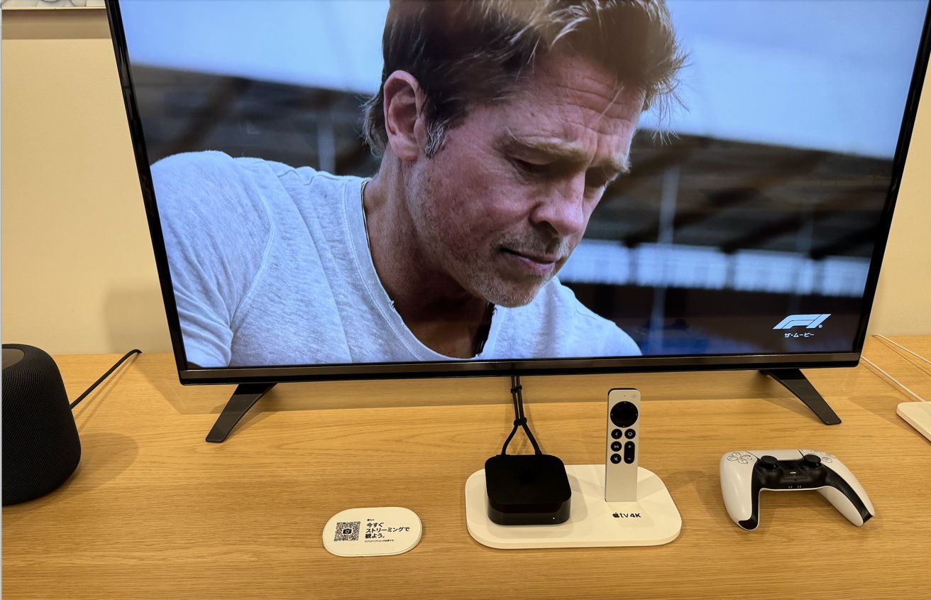 A TV screen displays a man in a white shirt. A black speaker, Apple TV box, remote, and video game controller are on a wooden table below.