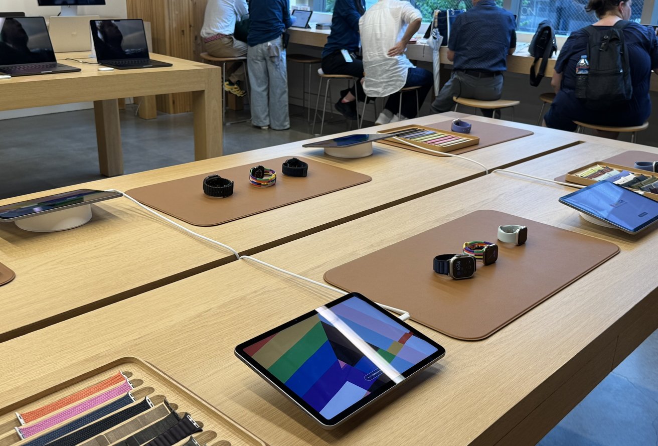 Retail display with smartwatches, colorful wristbands, and tablets on wooden tables. Customers seated at a counter using laptops in the background.