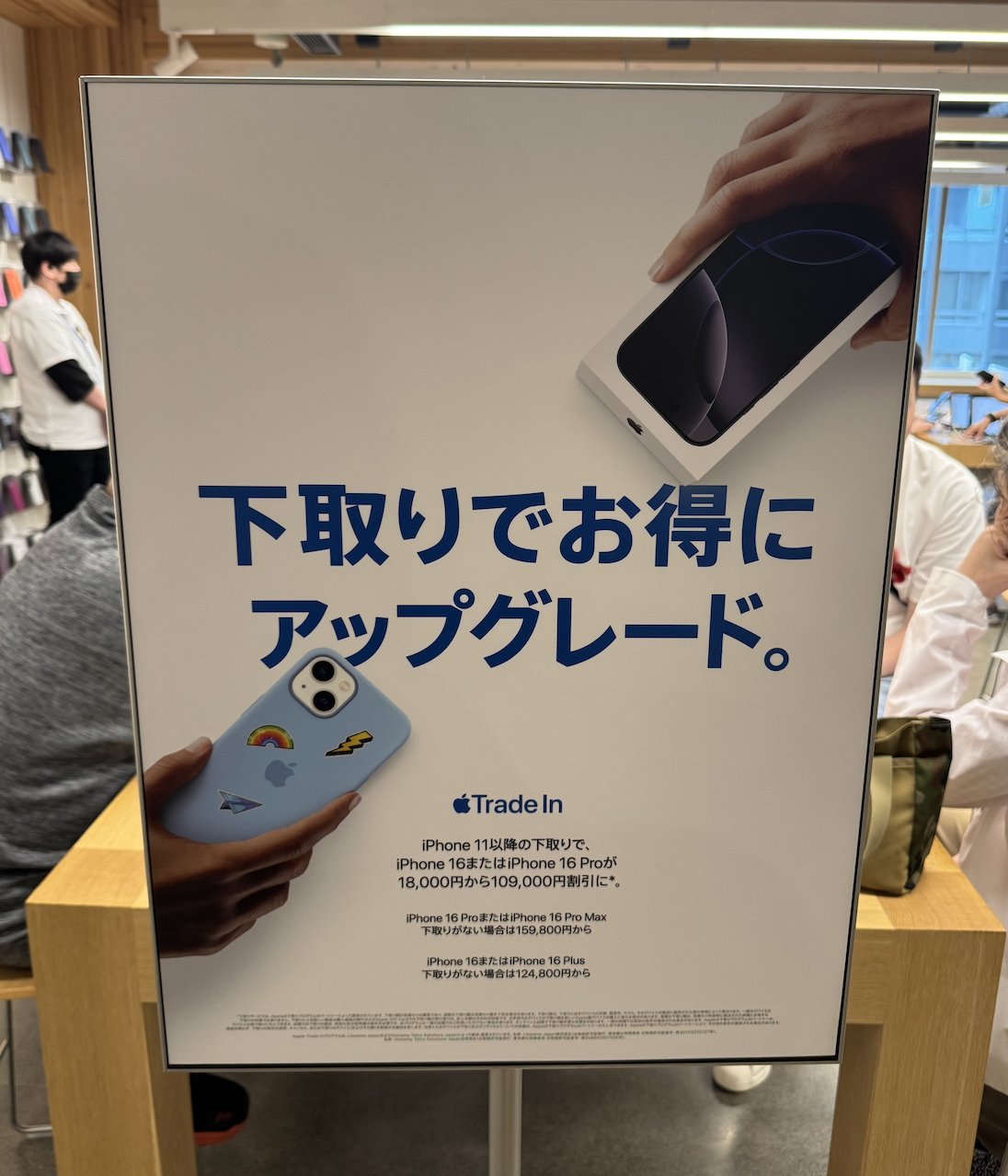 Promotional display for iPhone trade-in program featuring two hands holding iPhones. Japanese text and trade-in details visible in a retail store setting.