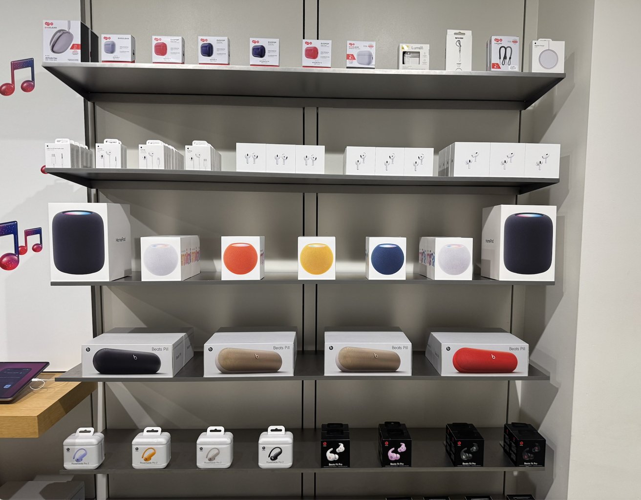 Store display with boxed smart speakers, earbuds, and headphones on shelves. Various brands and models are arranged neatly, mostly in white, grey, and black packaging.