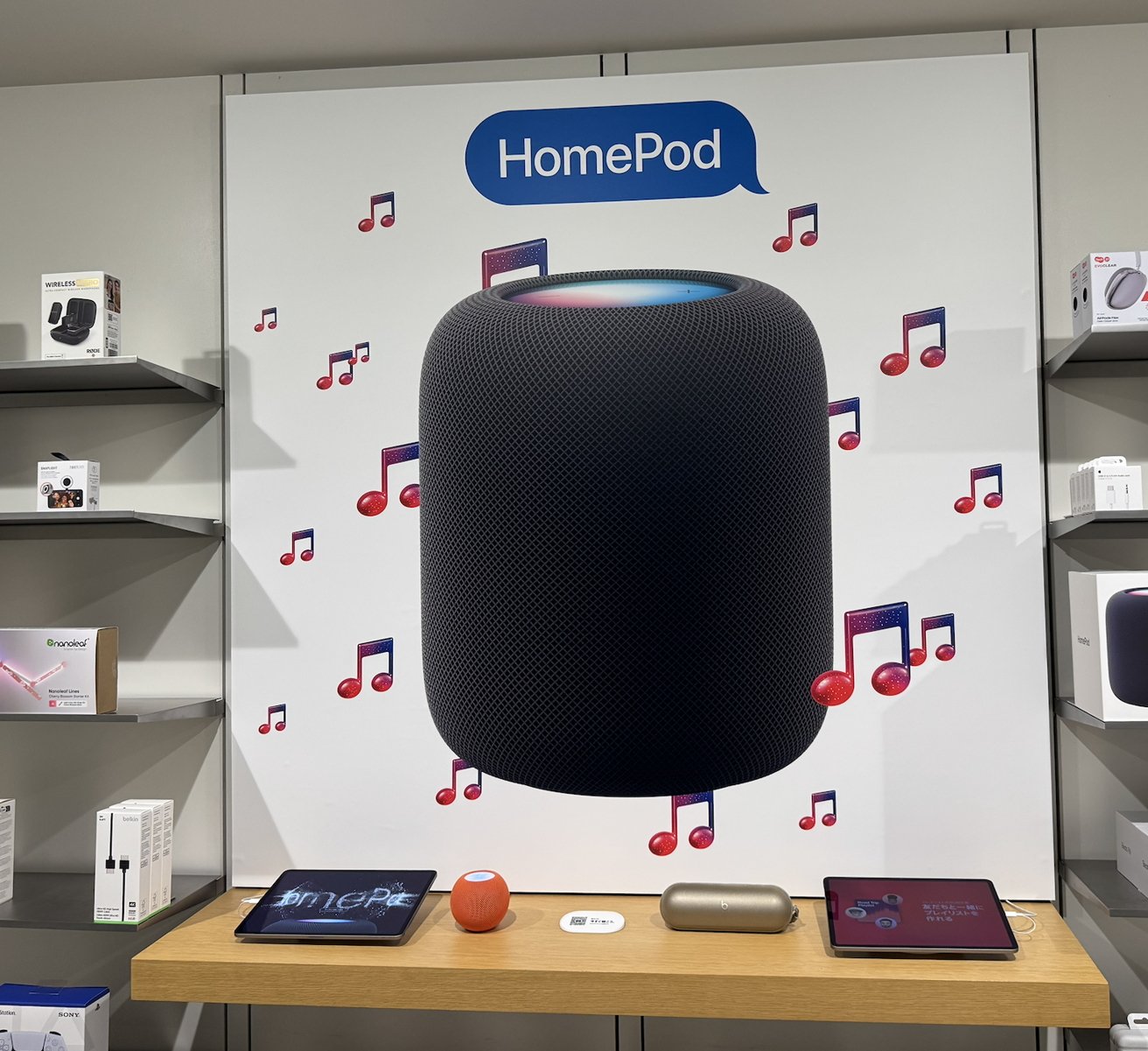 Large HomePod display with musical notes, surrounded by electronic devices on shelves and a table.