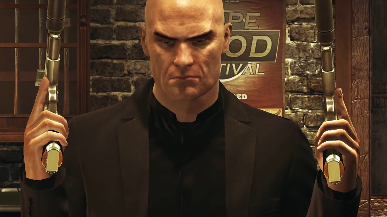 Bald man in a suit holds two guns, standing in front of a brick wall with a sign partially visible, serious expression on his face.
