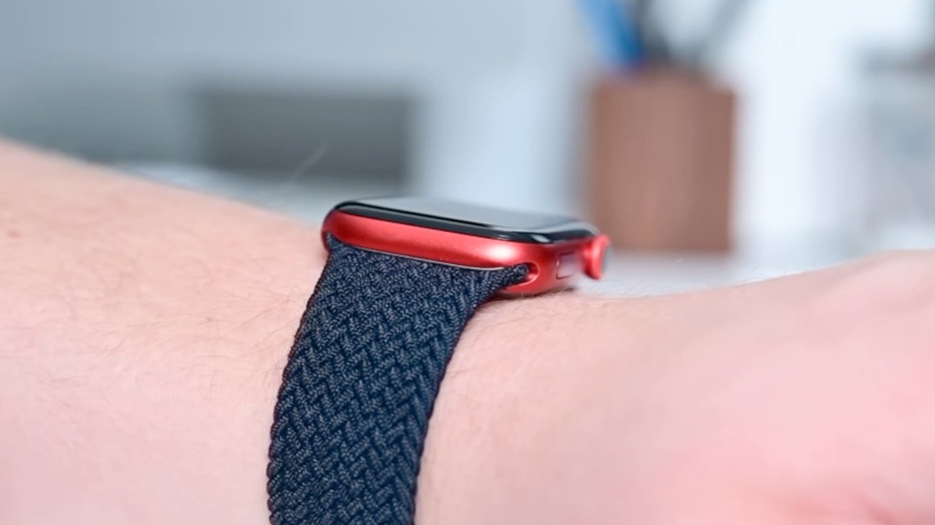 A red smartwatch with a blue woven band on a person's wrist, blurred background.