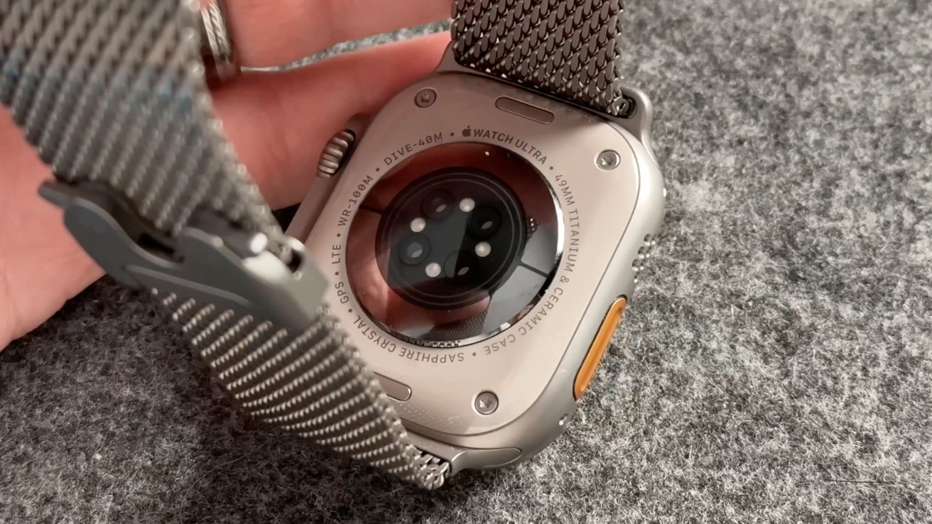Smartwatch back with sensors, titanium and ceramic case, sapphire crystal, text detailing watch specifications, and gray metal strap on gray fabric surface.