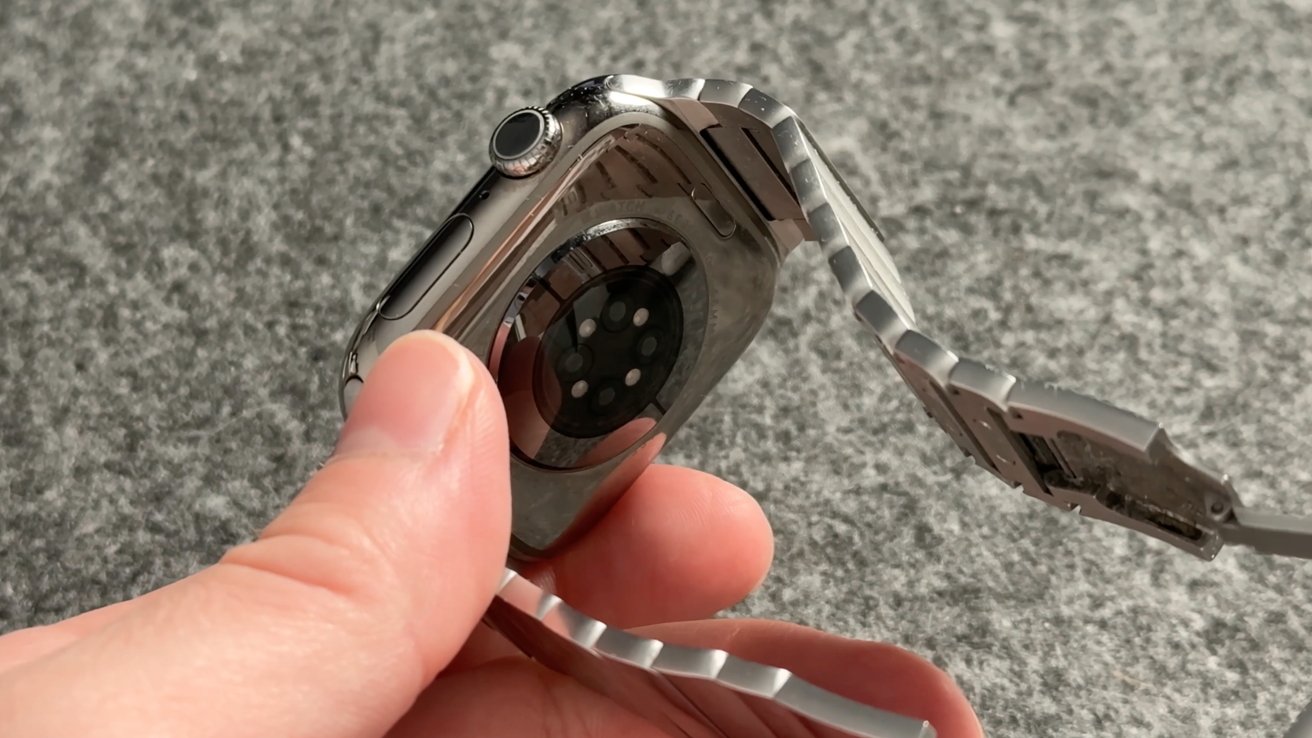 A hand holding a smartwatch with a metallic band, displaying the back sensor against a textured gray background.
