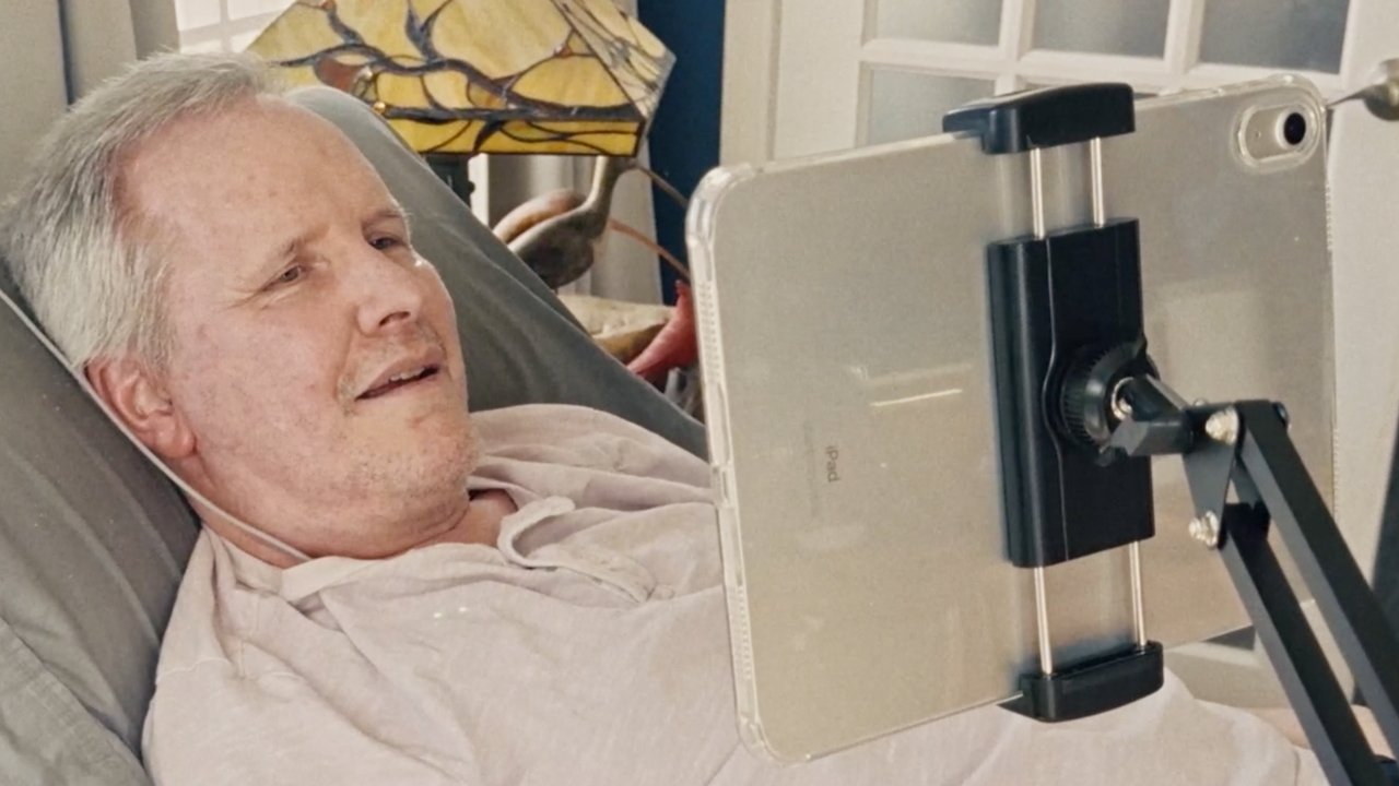Man lying in bed, watching tablet on adjustable stand, with warm lighting and a stained glass lamp in the background.