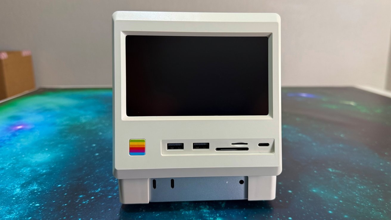Image: https://photos5.appleinsider.com/gallery/64602-134614-Wokyis-M5-review-10-xl.jpg A small white retro-style computer with a black screen and colorful logo on a blue-green space-themed surface