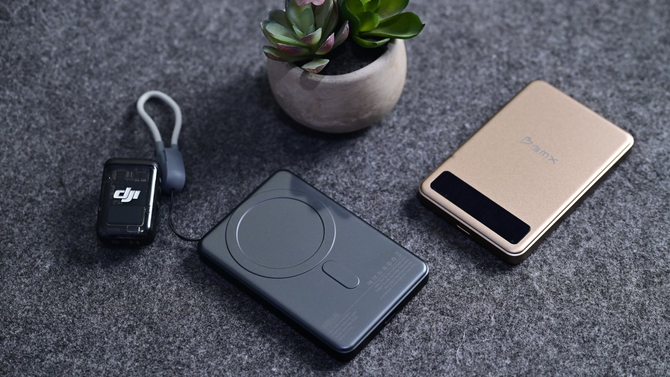 Tech gadgets and a small potted succulent on a gray surface; includes a black device with a strap, a gray wireless charger, and a gold power bank.
