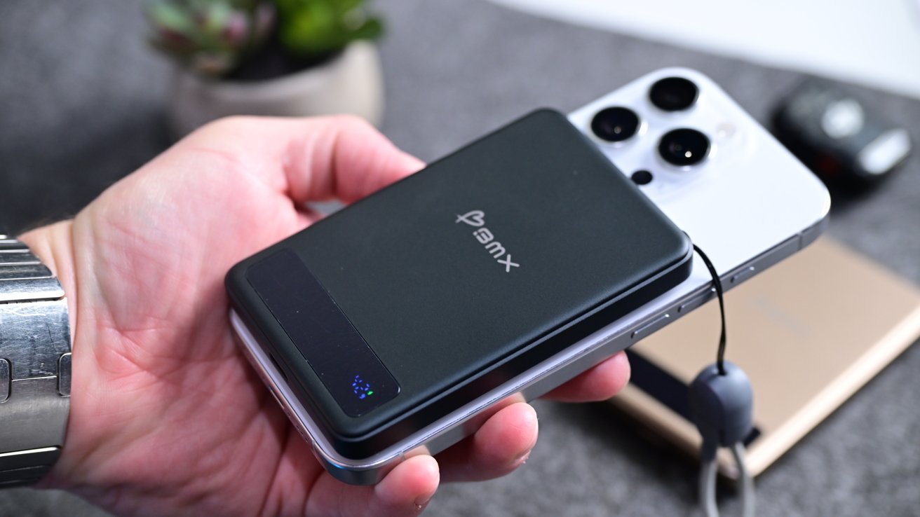 A hand holding a smartphone with a black power bank attached to the back, on a table with a plant and another device nearby.