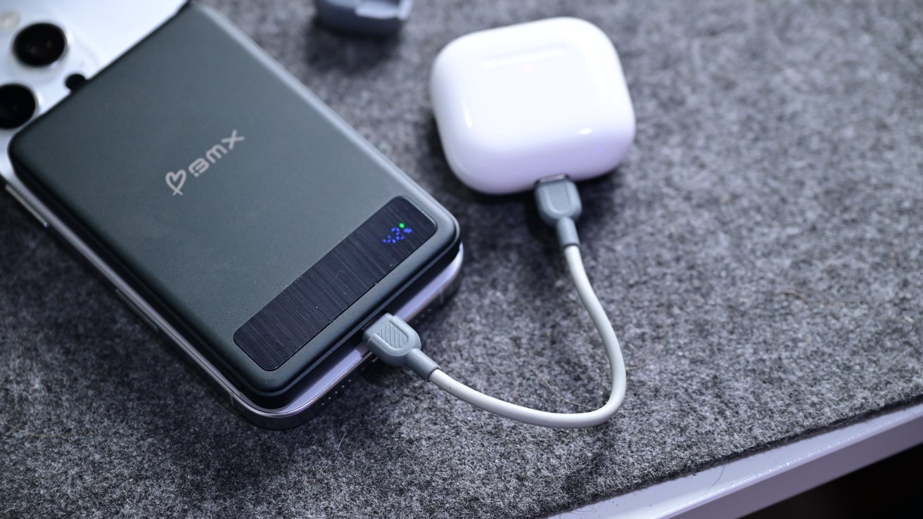 Smartphone with black case next to white wireless earbuds case, connected by a gray charging cable, on a textured gray surface.