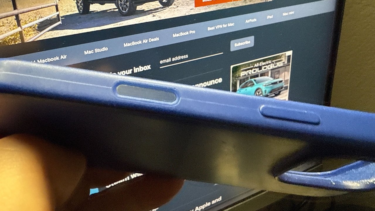 Close-up of a blue phone case's edge, revealing cutouts for buttons, with a computer screen displaying a website in the background.