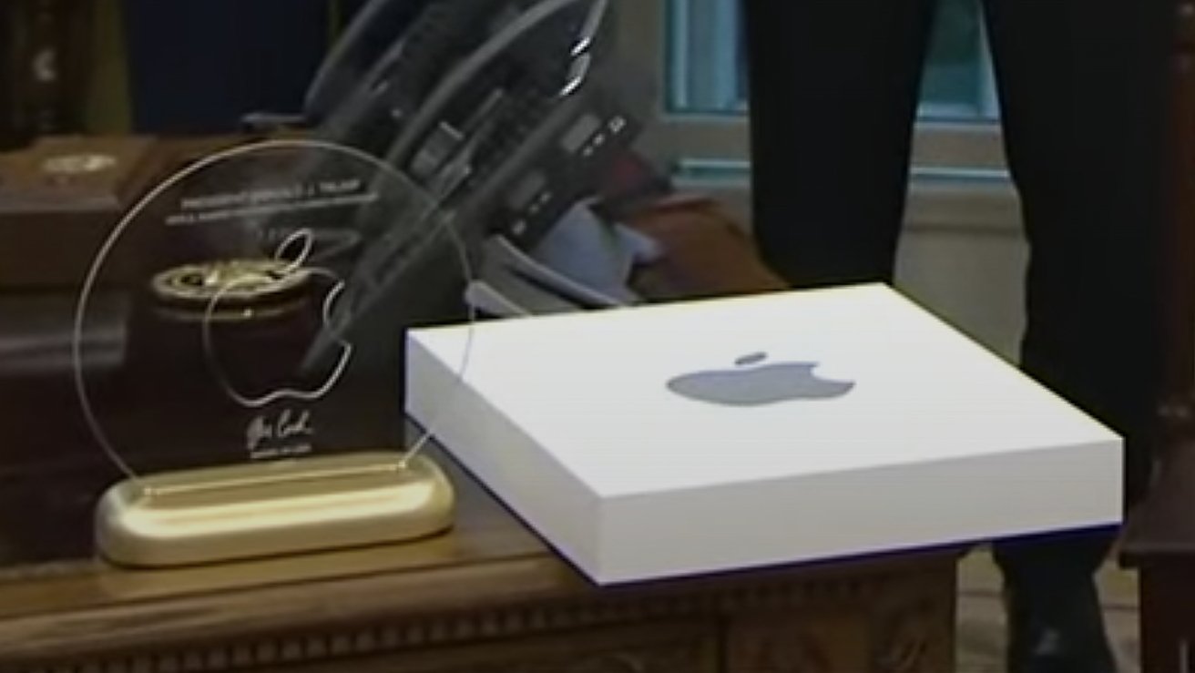 Glass plaque with Apple logo and name on a wooden table, beside a white box with Apple logo, telephones in background.