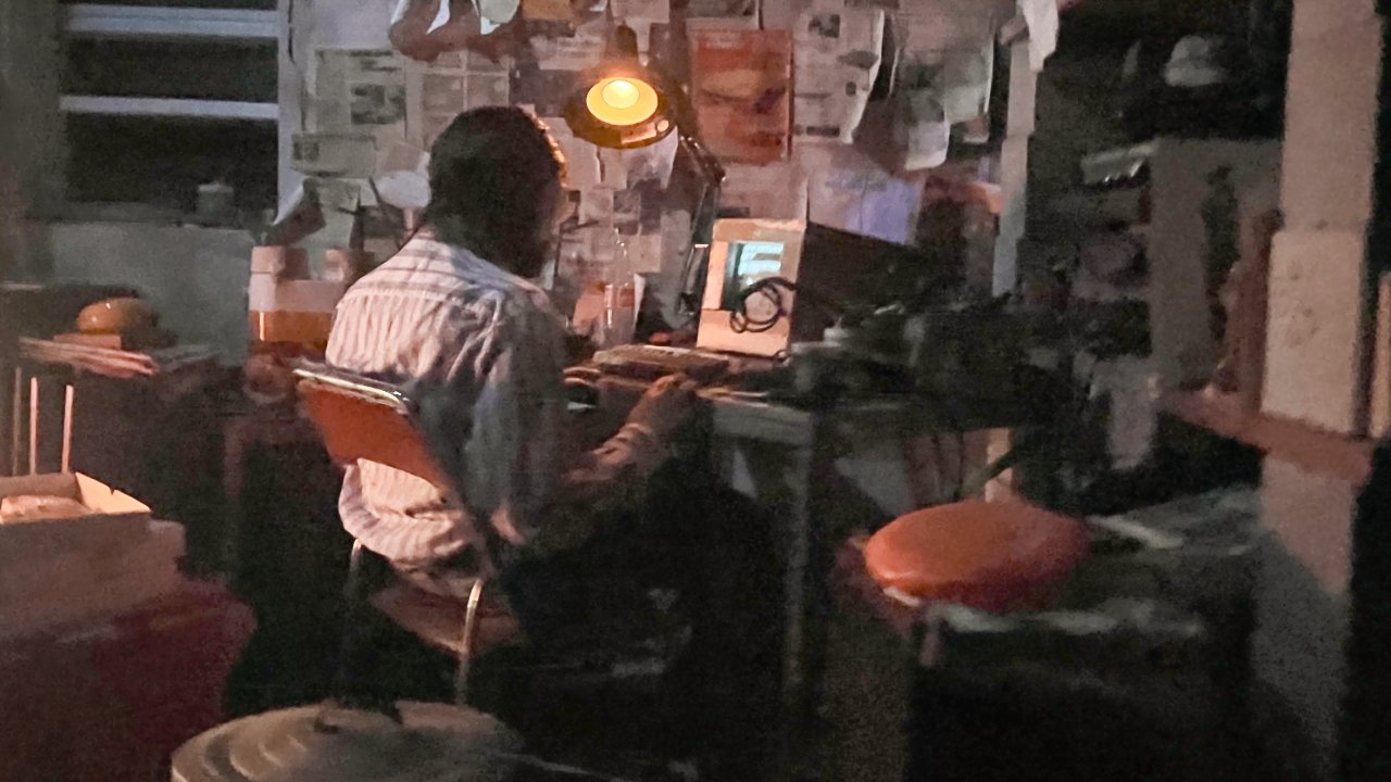 Person sitting at a cluttered desk, under a lamp, facing a computer. Wall covered with papers and notes in a dimly lit room.